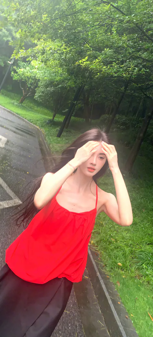 鞠婧祎身着红色吊带站在林间步道上，双手扶额，背景是葱郁绿树草地。