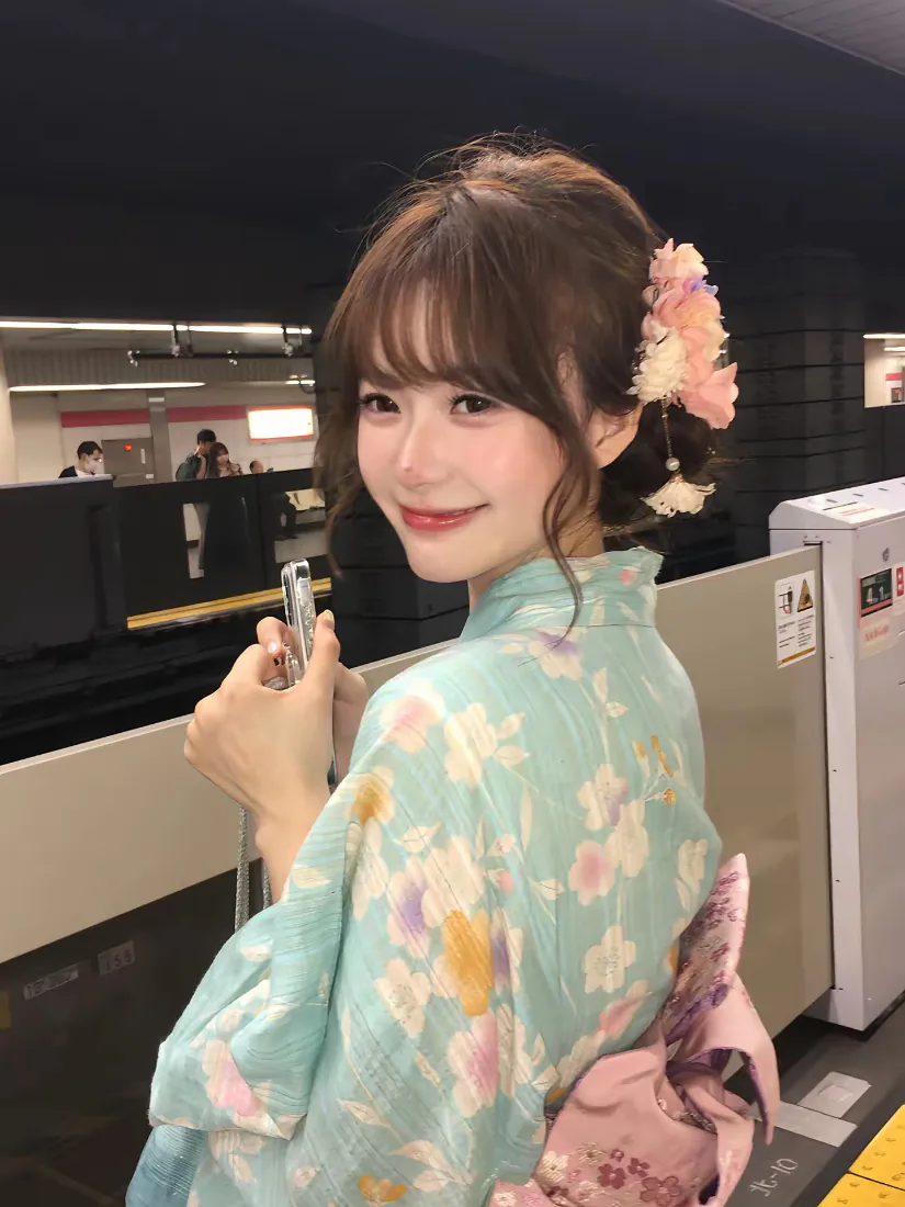 穿碎花和服的年轻女子在地铁站台回眸微笑，手持手机，发间别着樱花发饰。