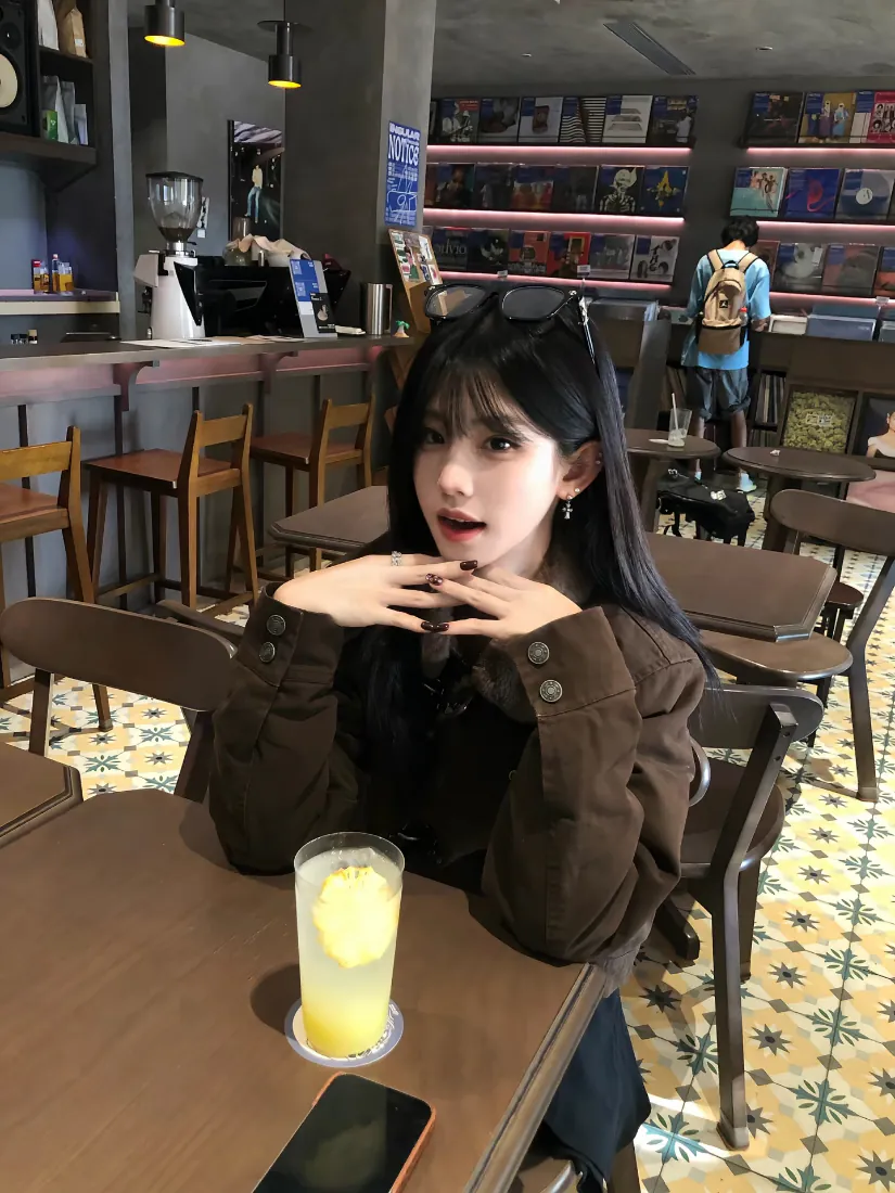 黑长直女生坐在咖啡馆桌前，桌上放着柠檬饮品与手机。