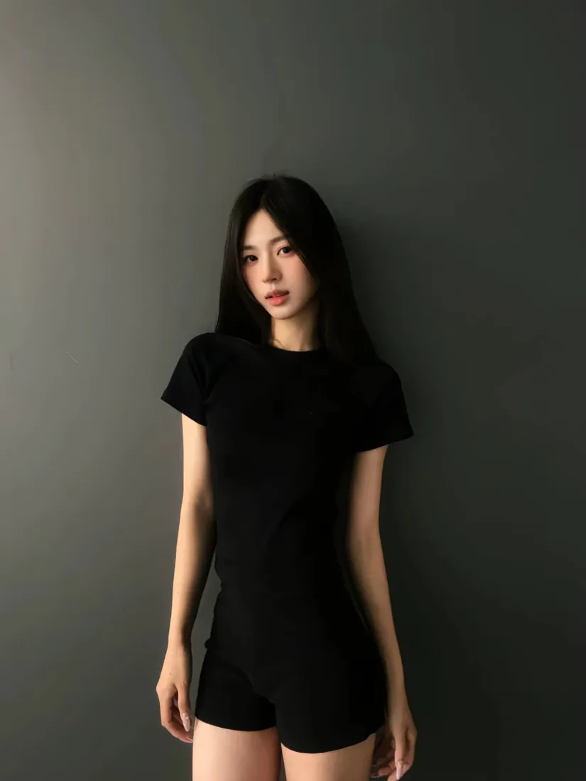 黑长直女生身着黑色短袖与短裤，倚靠在深灰色纯色墙面前。