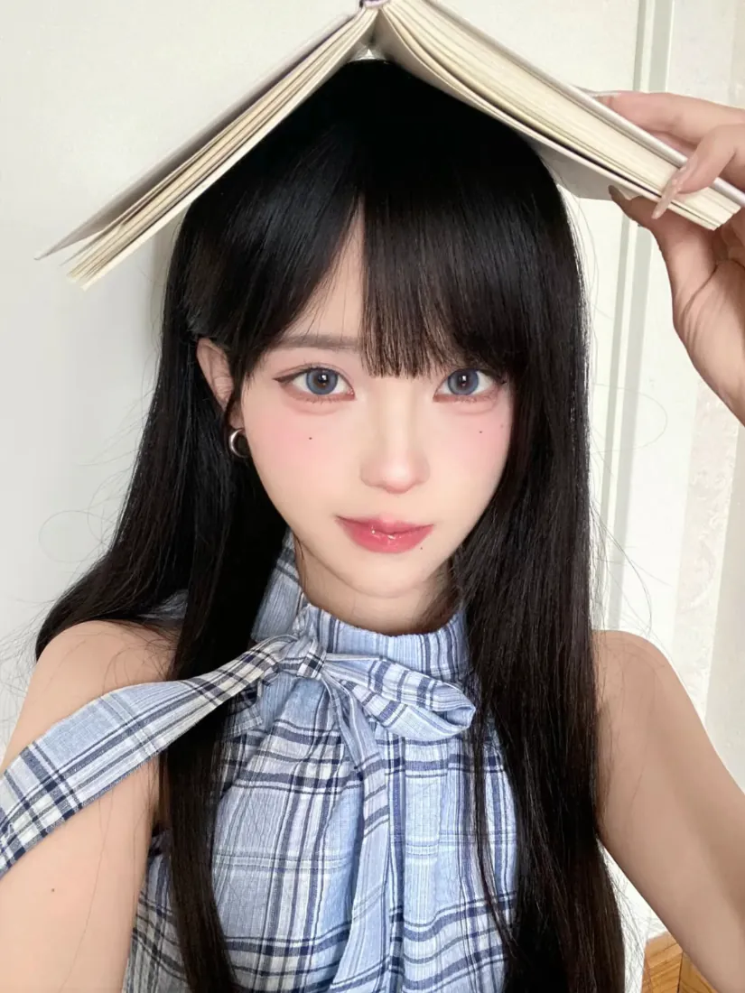 黑长直齐刘海女生穿蓝格纹挂脖上衣，将书本举在头顶做俏皮自拍。