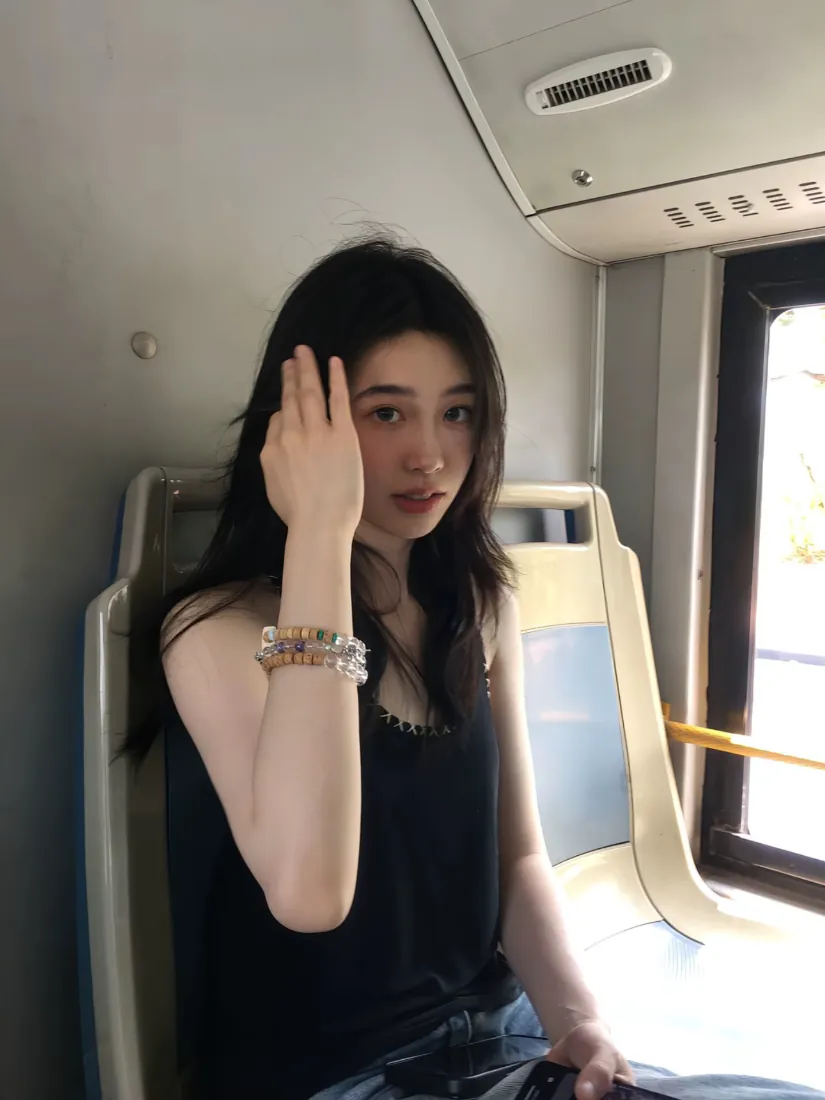 黑长发年轻女生坐在公交车内，穿黑色吊带，佩戴水晶手链，抬手轻触脸颊。