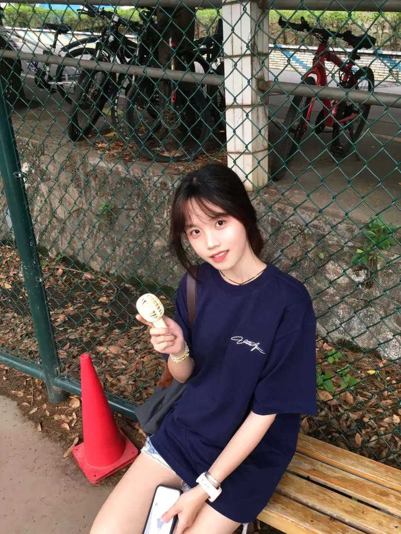 年轻少女坐在户外木长椅上，手持雪糕，身后是铁丝网围栏与停放的自行车。