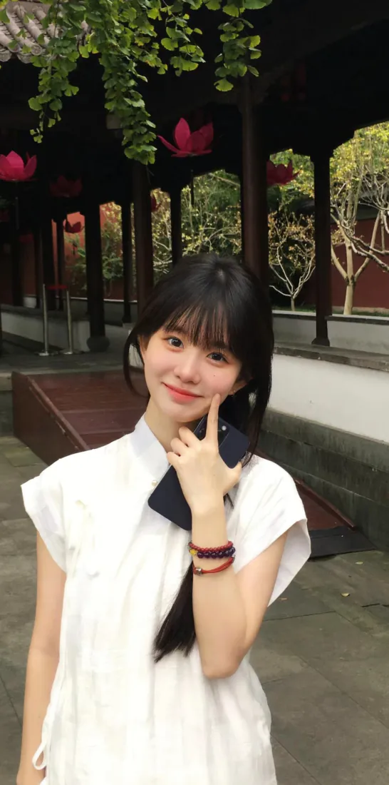 白衣齐刘海少女在中式古建庭院前比耶微笑自拍，背景有绿植与红色装饰。