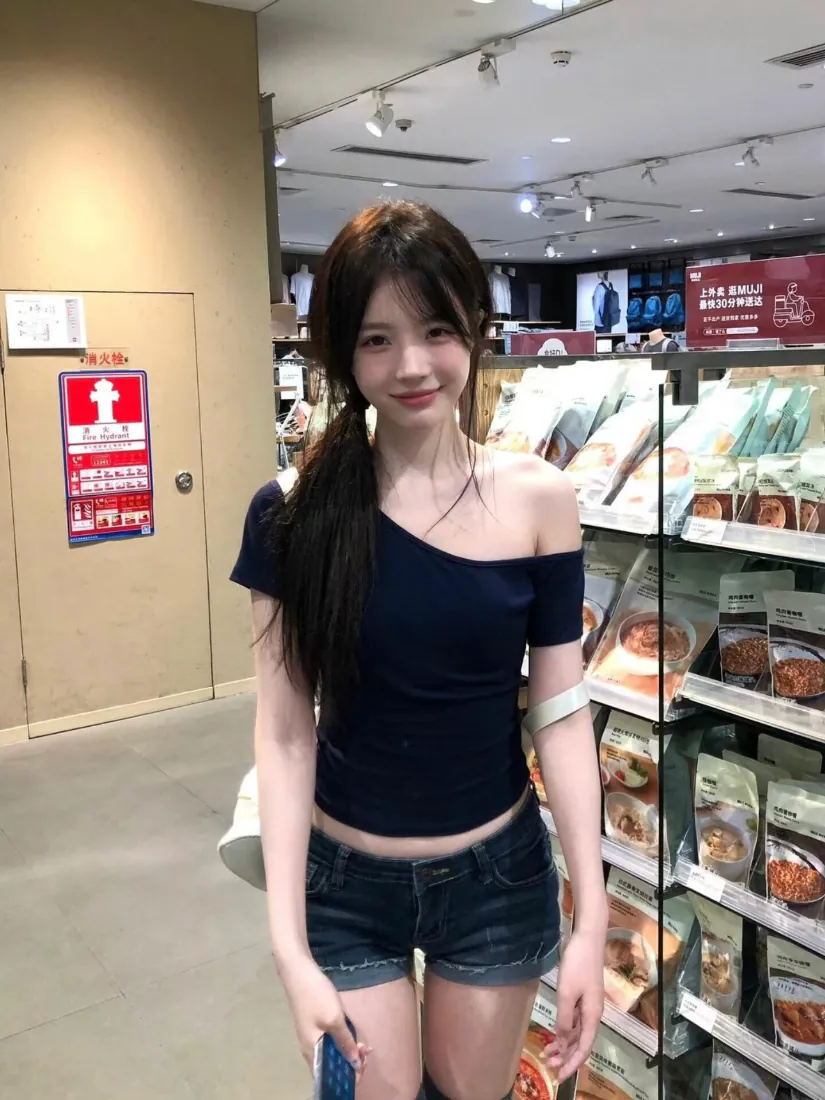 长发年轻女子身着藏蓝斜肩露腰上衣与牛仔短裤，在超市货架前微笑自拍。