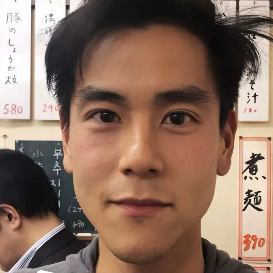 彭于晏在拉面店内正对镜头自拍，背景墙面挂有日文餐牌，左侧有背对镜头的男子。