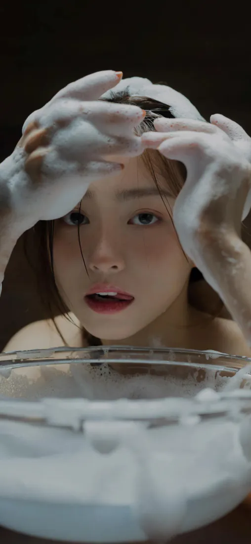 暗调背景下绿瞳少女双手沾满洗发泡沫，正低头揉搓头发，身前摆放盛有泡沫的透明容器。