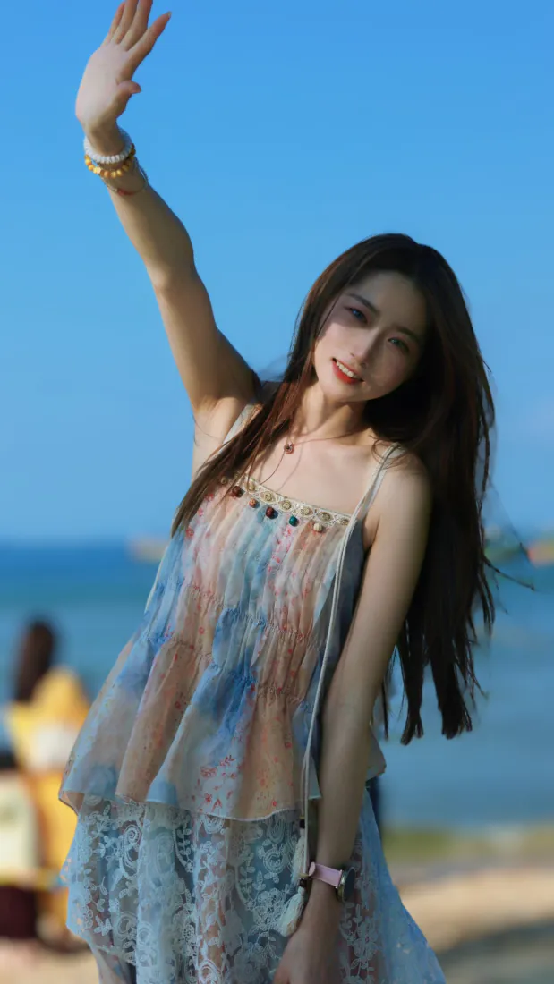 长发少女身着渐变吊带裙在海边沙滩抬手挥手，背景是澄澈蓝天与蓝海。