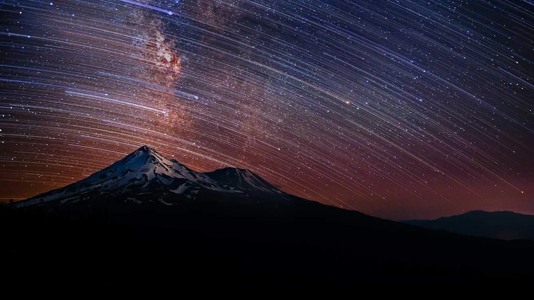 2k星空壁纸|雪山银河星轨夜景背景图片 - 壮阔震撼风光「哲风壁纸」 夜晚雪山剪影上方,布满斜向星轨的银河夜空,天际晕染橙红渐变