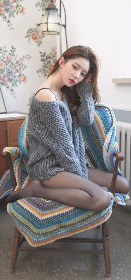 长发美女穿灰色露肩毛衣黑丝，跪坐于铺彩色针织毯的木椅上，居家碎花墙前。