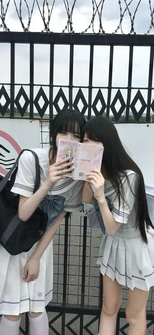 两名穿JK制服的黑发少女在铁丝网围栏前，共举粉色手账本遮挡面部。